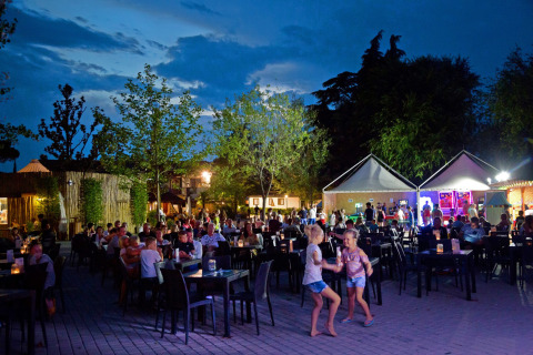 Tarde en Altomincio Family Park en Lombardía con familias cenando, niños jugando y luces tenues al aire libre.