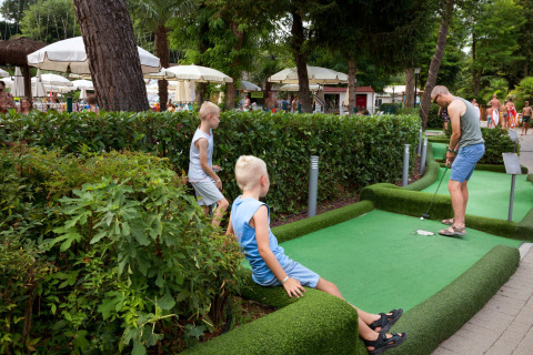 Familia jugando al mini golf en Altomincio Family Park en Lombardía, Italia, rodeados de vegetación y sombrillas.