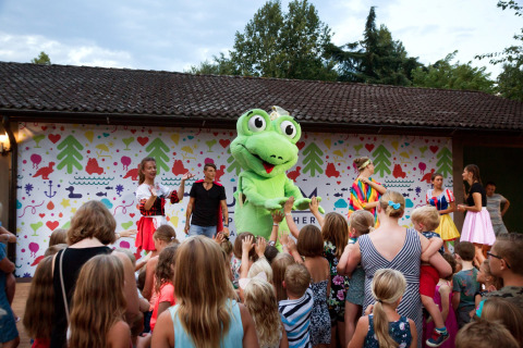 Niños se reúnen frente a un escenario con una mascota verde y adultos disfrazados en Altomincio Family Park, Lombardía.