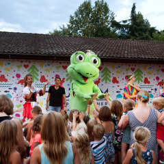 Niños se reúnen frente a un escenario con una mascota verde y adultos disfrazados en Altomincio Family Park, Lombardía.