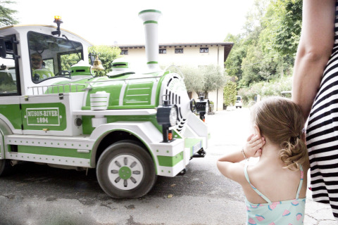 Niña pequeña se tapa los oídos mientras pasa un tren verde y blanco en Altomincio Family Park, Italia.