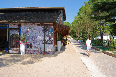 Sti ved Altomincio Family Park i Lombardiet, Italien, med folk der går langs en bygning omgivet af træer.