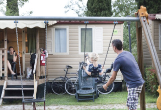 Een man duwt een kind op een schommel voor een stacaravan, terwijl een vrouw op het terras zit.