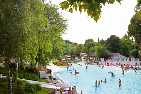 Parque vacacional en Lombardía, Italia, con gran piscina, familias, niños y rodeado de vegetación verde.