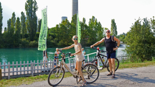 Dos personas con bicicletas junto al río en Altomincio Family Park, Lombardía, Italia, en un día soleado.