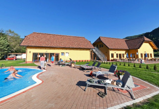 Camping Bella Austria nel parco vacanze in Stiria, Austria, con edifici gialli, piscina, lettini e ospiti rilassati.