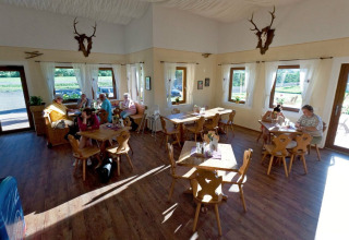 Indoor café at Camping Bella Austria, featuring wooden tables, guests, large windows, and scenic outdoor views.