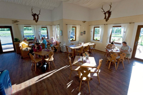 Café interior en Camping Bella Austria con mesas de madera, huéspedes disfrutando y ventanas con vistas verdes.