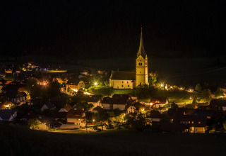 Foto notturna di Camping Bella Austria in Stiria, Austria, con case illuminate e una chiesa centrale.