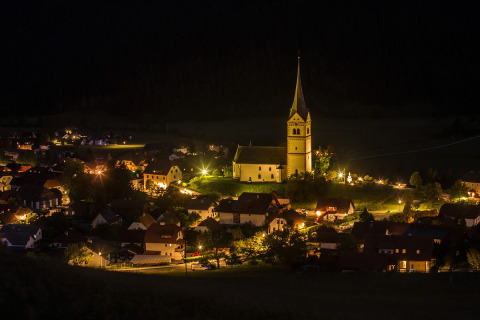 Foto notturna di Camping Bella Austria in Stiria, Austria, con case illuminate e una chiesa centrale.