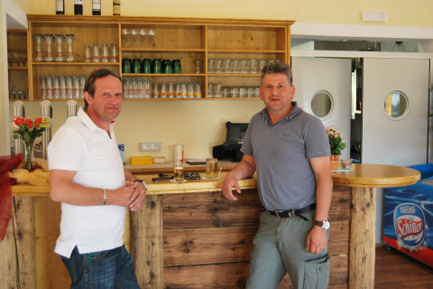 Due uomini al bancone del bar presso Camping Bella Austria, Stiria, Austria, circondati da bicchieri e bevande.