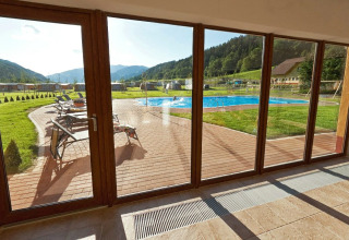 Vista a través de grandes ventanales al área de la piscina y colinas en Camping Bella Austria en Estiria.