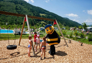 Children play on a playground with swings, joined by a bee mascot, near a pool and green mountains.