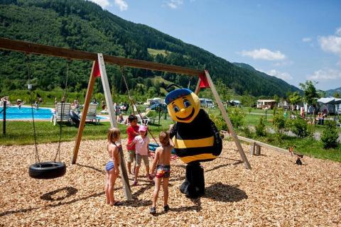 Children play on a playground with swings, joined by a bee mascot, near a pool and green mountains.