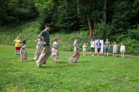 Children enjoy a sack race on a green field at Camping Bella Austria, with spectators and forest behind.