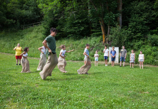 Children enjoy a sack race on a green field at Camping Bella Austria, with spectators and forest behind.