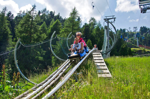 Two people ride a mountain coaster at Camping Bella Austria, surrounded by green hills and forest trees.