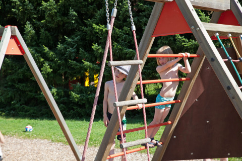 Niños con bañadores juegan en una estructura de escalada en Camping Bella Austria, rodeados de árboles verdes.
