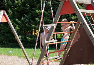 Kinderen in zwemkleding spelen op een klimrek bij Camping Bella Austria, omringd door groene bomen.