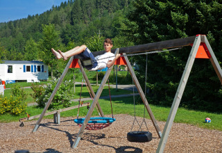 Ragazzo gioca sull’altalena al Camping Bella Austria in Stiria, con alberi verdi e roulotte sullo sfondo.