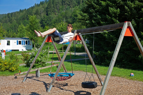 Jongen speelt op een schommel bij Camping Bella Austria, Stiermarken, met bomen en caravans op de achtergrond.