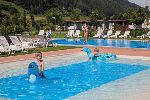 Niños juegan en una piscina poco profunda con inflables en Camping Bella Austria, rodeados de tumbonas y cabañas.