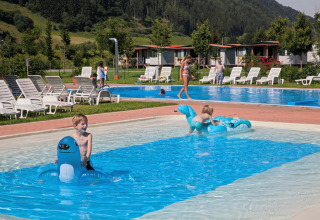 Børn leger i en lav swimmingpool med oppustelige dyr på Camping Bella Austria, omringet af liggestole og hytter.