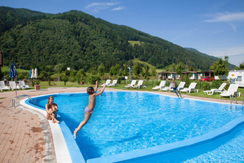 Niños saltando a la piscina rodeada de tumbonas y montañas en Camping Bella Austria, Estiria, Austria.