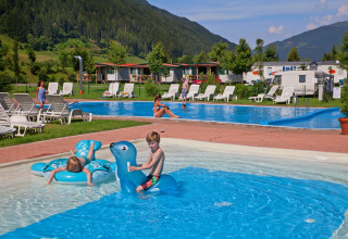Børn leger i en pool med oppustelige dyr på Camping Bella Austria, omgivet af bjerge og feriehytter.