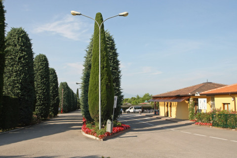 Entrada a Camping Bella Italia en Véneto, Italia, rodeada de cipreses y un edificio amarillo bajo el sol.