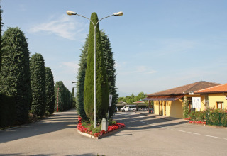 Ingresso del Camping Bella Italia in Veneto, Italia, fiancheggiato da cipressi e un edificio giallo al sole.