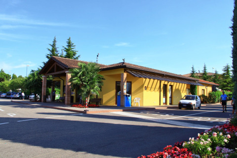 Recepción del Camping Bella Italia en Véneto, Italia, con edificio amarillo, autos y paisaje ajardinado.