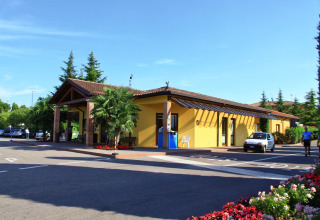 Reception del Camping Bella Italia, parco vacanze in Veneto, Italia, con edificio giallo e auto parcheggiate.
