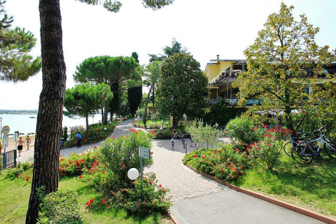 Sentiero tra fiori e alberi a Camping Bella Italia, villaggio turistico in Veneto, Italia, vicino al lago e agli edifici.