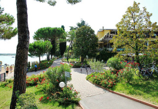 Sti omgivet af blomster og træer på Camping Bella Italia, en feriepark i Veneto, Italien, nær sø og bygninger.