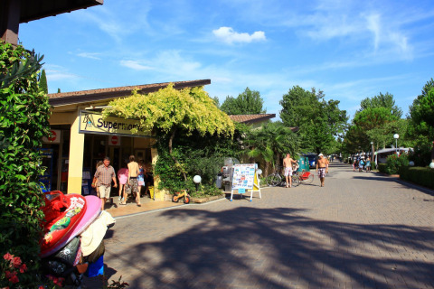 Día soleado en Camping Bella Italia, Véneto, Italia, con personas entrando al supermercado entre árboles verdes.