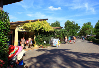 Sunny day at Camping Bella Italia, Veneto, Italy, with people walking by a supermarket and lush greenery.