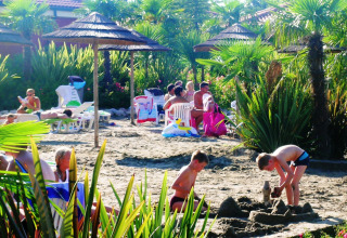 Bambini che giocano sulla sabbia tra palme e ombrelloni, mentre le famiglie si rilassano al Camping Bella Italia.
