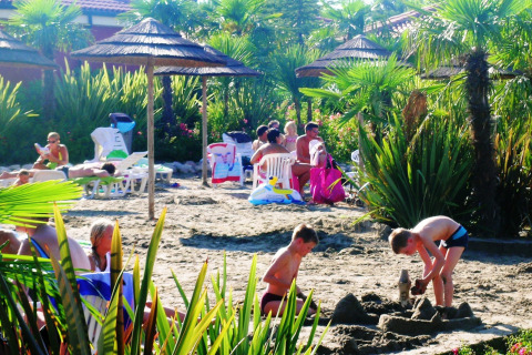 Children play in the sand at a palm-lined beach with parasols, while families relax at Camping Bella Italia in Italy.