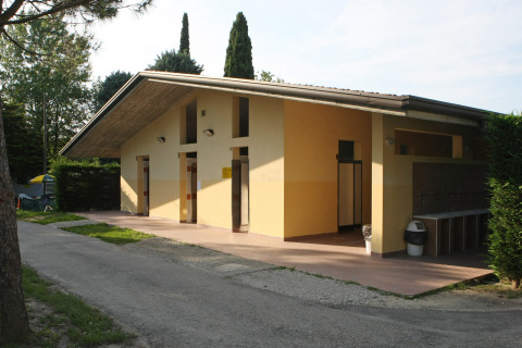 Edificio sanitario exterior en Camping Bella Italia, parque vacacional en Véneto, Italia, rodeado de árboles.