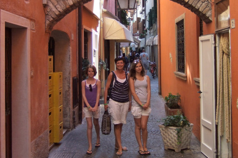 Drie vrouwen wandelen door een smalle, kleurrijke steeg in Camping Bella Italia, Veneto, Italië, omringd door gebouwen.