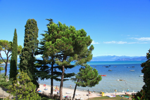 Playa y árboles en Camping Bella Italia en Véneto, Italia, con vistas al lago y visitantes tomando el sol.