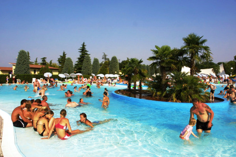 Famiglie e bambini si divertono nella piscina all’aperto tra palme al Camping Bella Italia, Veneto, Italia.