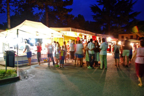 Evening photo of a lively market at Camping Bella Italia in Veneto, Italy, with people and illuminated stalls.