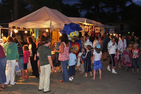Mercatino serale al Camping Bella Italia in Italia con famiglie, bambini e bancarelle sotto tende illuminate.