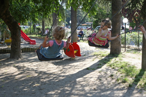 Dos niños juegan en columpios en un parque infantil sombreado en Camping Bella Italia, Véneto, Italia.