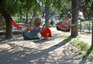 Due bambine giocano sulle altalene in un’area giochi ombreggiata al Camping Bella Italia, Veneto.