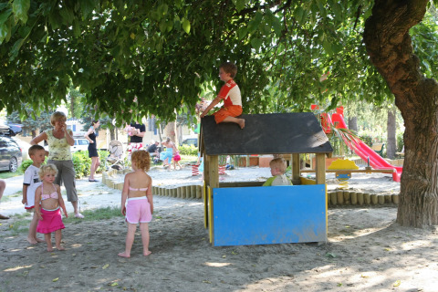 Children play under a large tree at the playground in Camping Bella Italia, Veneto, Italy, with adults nearby.