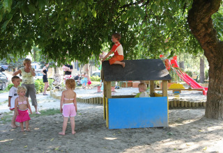 Bambini giocano sotto un grande albero nell’area giochi di Camping Bella Italia, Veneto, Italia, con adulti vicini.
