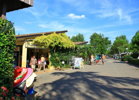 Guests walk past the supermarket at Camping Bella Italia, surrounded by trees in Veneto, Italy.